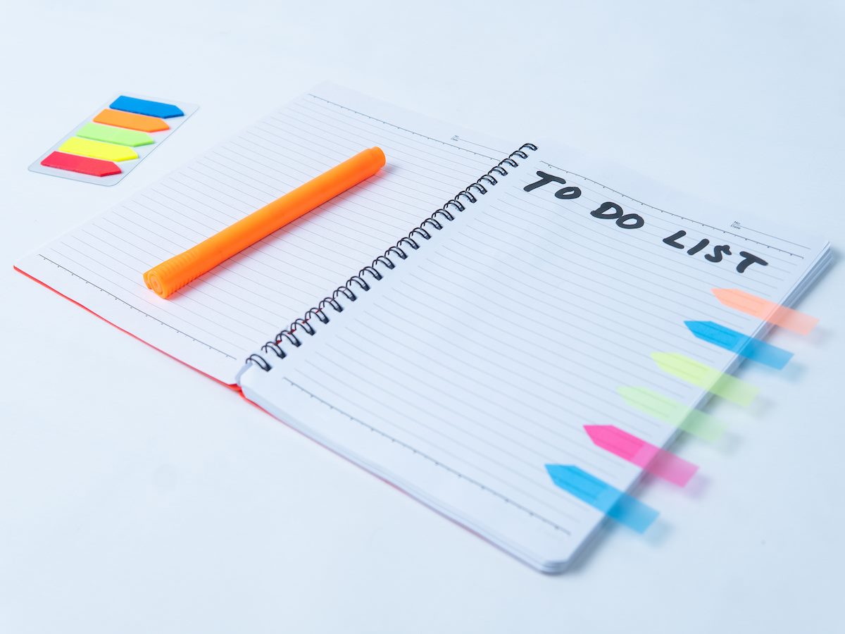 an open spiral notebook with handwritten to do list, orange pen, and colorful sticky notes on white background