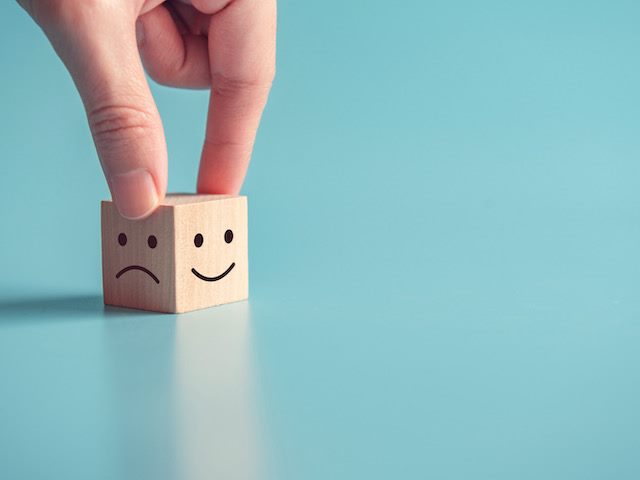 fingers on a cube with one side showing a happy face and the other side showing a sad
