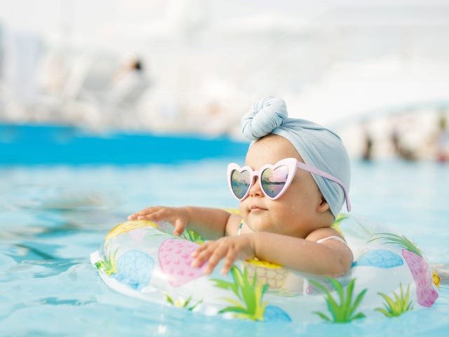 Toddler girl with knotted headscarf and in colorful swimsuit and heart-shaped sunglasses relaxing on inflatable toy ring floating in pool have fun during summer vacation in tropical resort.