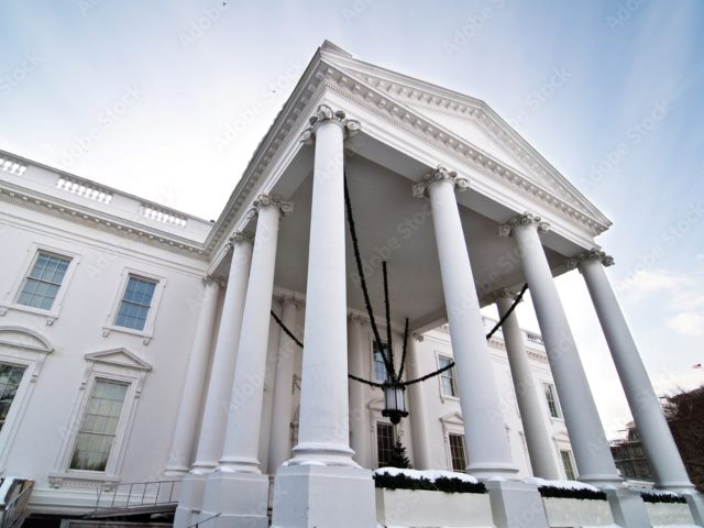 An angled shot of the facade of The White House emphasizing it's columns and lighting in reported story of President Biden requiring pledge from AI companies on safety and security.
