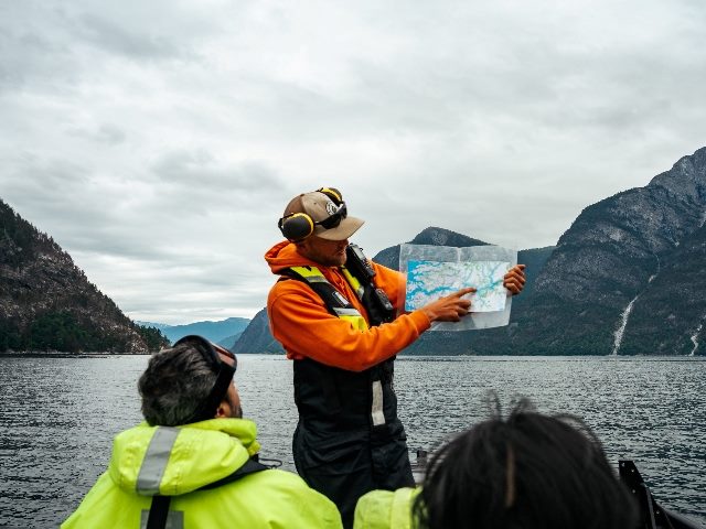 People on a boat getting a fjord tour. Shows the back side of two people sitting in boat listening to tour guide who is standing point to a map in Sognefjord, Norway.