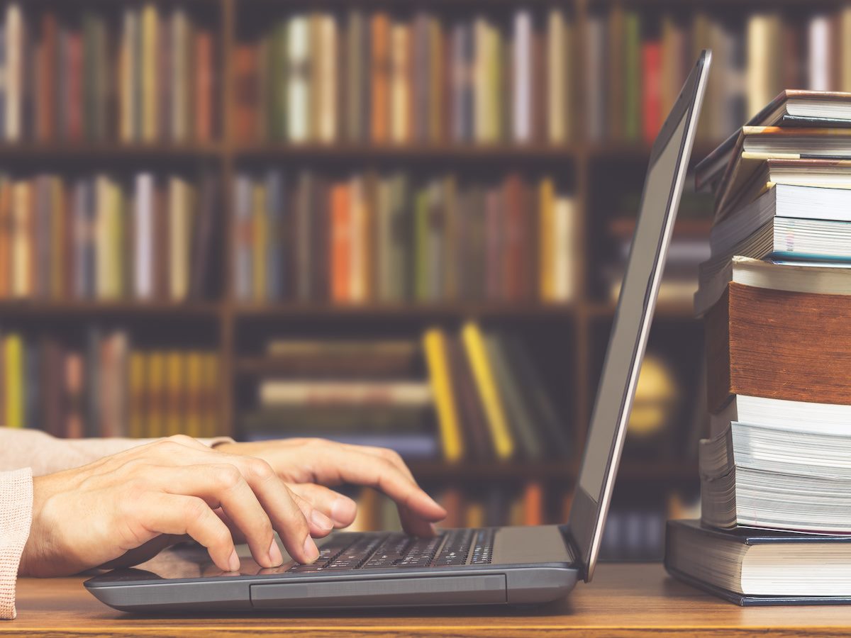 Hands typing on a laptop with a stack of books behind it 