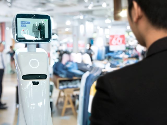 A robotic assistant in retail store to help customers navigate to items of interest. The assistant is a white column with a screen attached to the top and a man in a black suit is in the foreground with his back to the camera and appears to be perusing the assistant's content with the store blurred in the background. 
