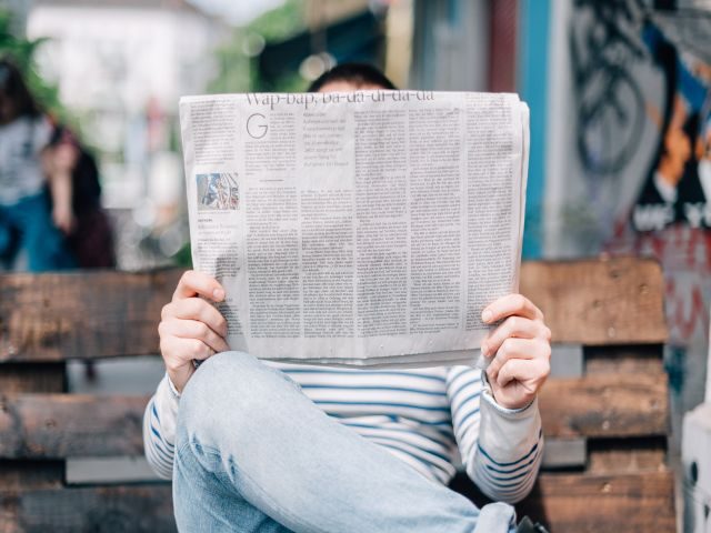 A man reading a newspaper, while sitting on a park bench