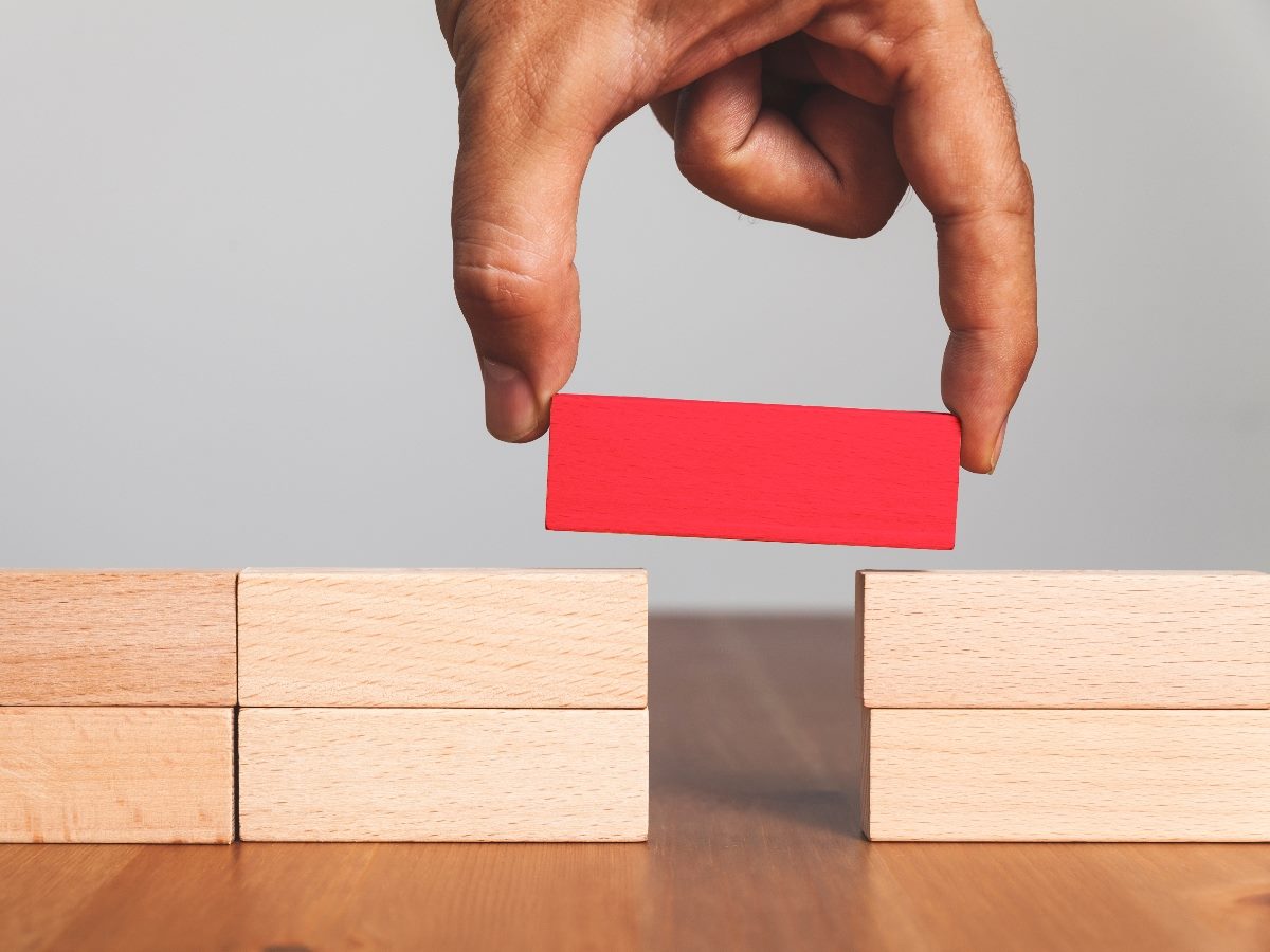 A hand places a red wooden block between two stacks of beige blocks, symbolizing bridging a gap or connecting missing pieces.
