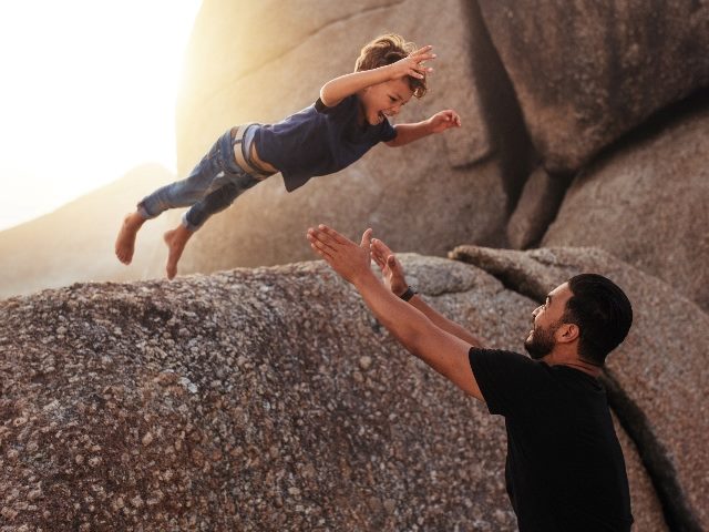 A young boy jumps from a large rock on a beach into the arms of an adult male standing waiting to catch him with golden sunshine in the background, suggesting the importance of trust in CX and EX.