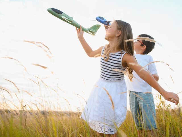 A boy and a girl stand in a sunlit field each holding and about to throw a toy airplane in the same direction (to the left).