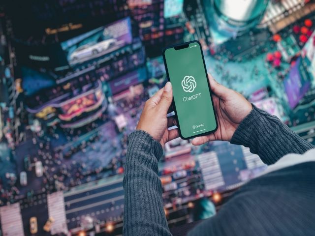 Girl on balcony of high floor apartment holding smartphone with ChatGPT artificial intelligence chatbot app on screen. City at night in the background. 