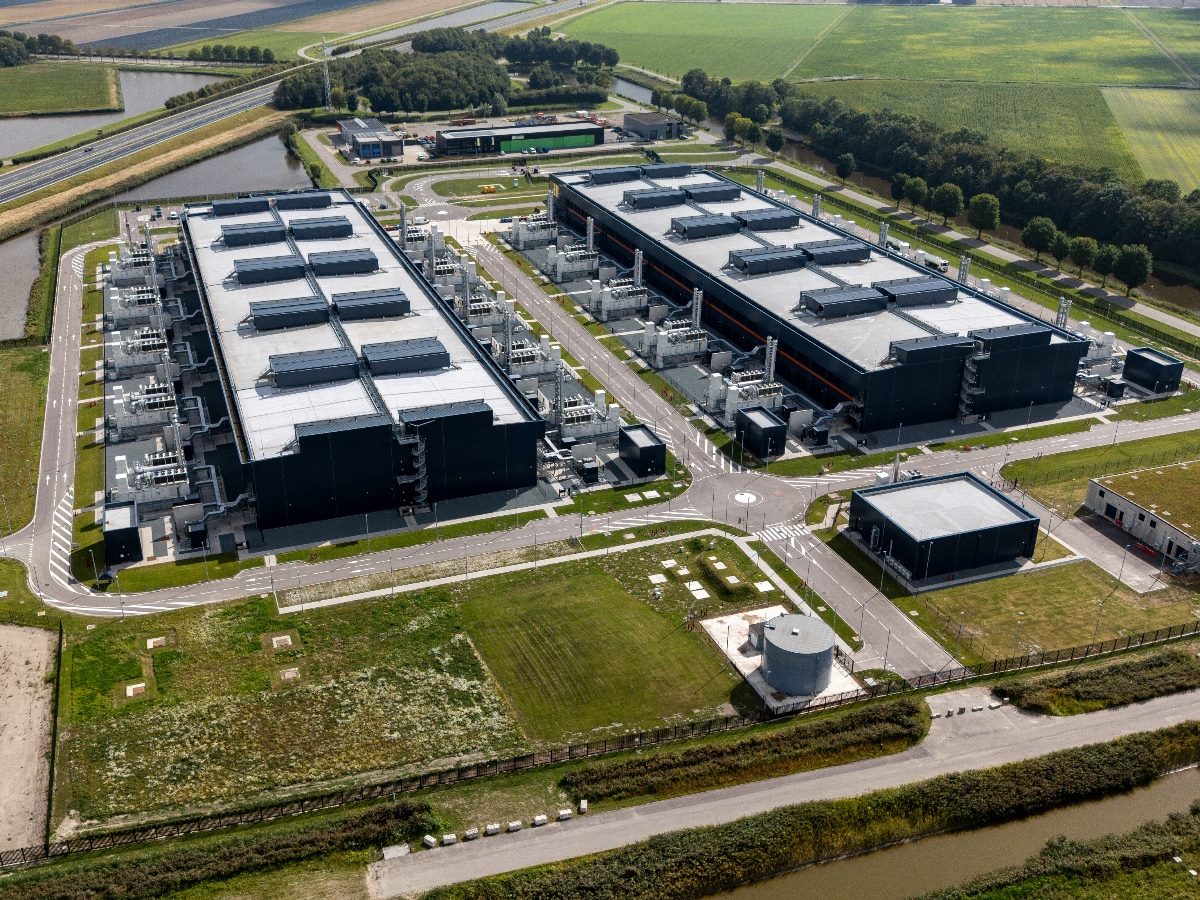 Aerial view of a large modern data center campus with two massive rectangular server buildings, cooling and power equipment lining the sides, internal roads, security fencing, canals and surrounding green farmland.