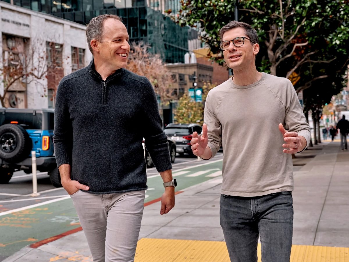 Bret Taylor and Clay Bavor walk side by side on a city sidewalk, talking and gesturing during a conversation outside in an urban setting.