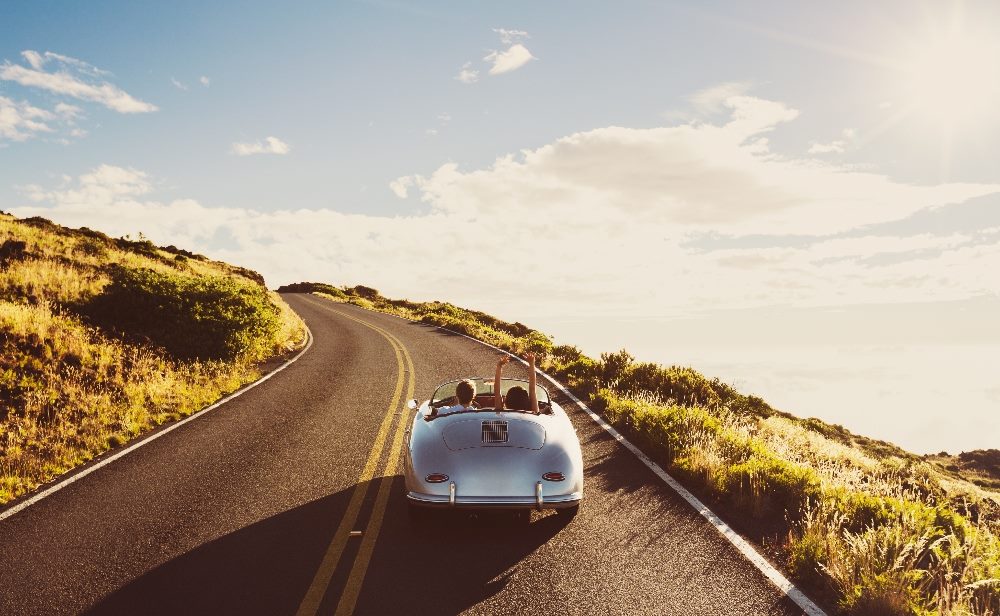 A silver vintage coupe with a couple inside with the woman raising her arms into the air drives down a beachside road, suggesting the benefits of generative AI in customer experience on the customer journey.