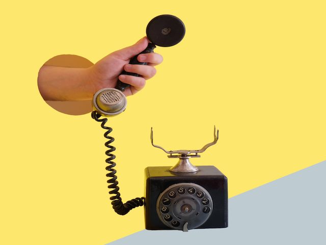 surreal image of arm coming out of yellow wall holding the receiver of an old phone
