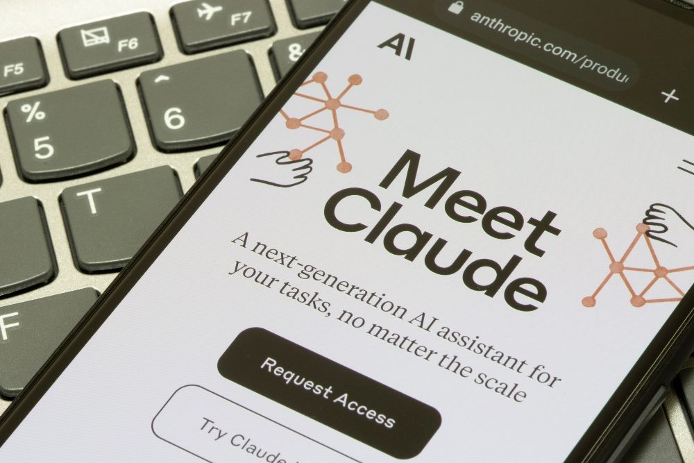 A smartphone screen displays the "Meet Claude" page from Anthropic's website. The page describes Claude as a next-generation AI assistant for various tasks. The screen shows a "Request Access" button and features simple illustrations of network connections. The smartphone is placed on a keyboard, highlighting the technology context in piece about the new Claude upgrade, Claude 3.5 Sonnet.