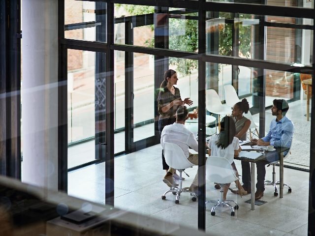 A woman leading her team through a meeting behind a glass wall. Four employees sitting at table listen in.