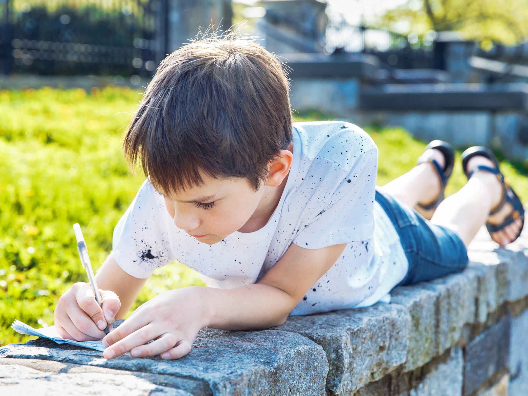 A boy wearing a white T-shirt, shorts, and sandals is lying on a stone fence, writing in a notepad.