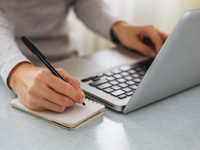 A close-up of a person seated at a laptop computer, their left hand on the keyboard and their right hand taking notes with a pen and notepad