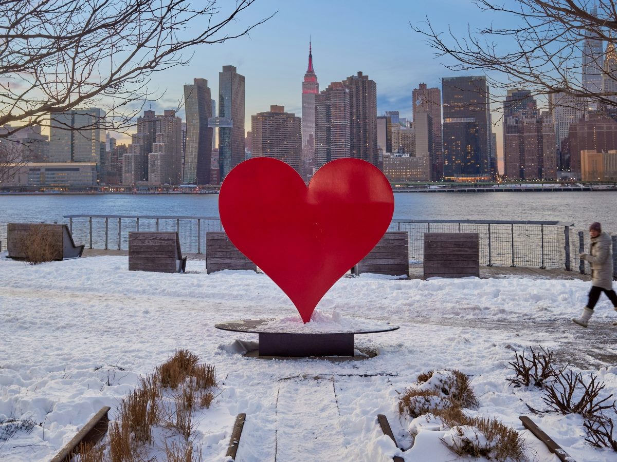 Heart monument in a snowy scene with a river and city skyline in the background.