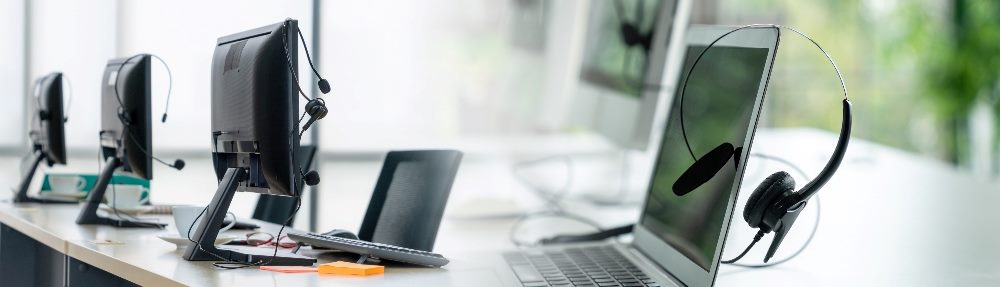 Laptops sit on a long work table with headsets draped over their screens in piece about the impact of AI in contact centers on the workforce.
