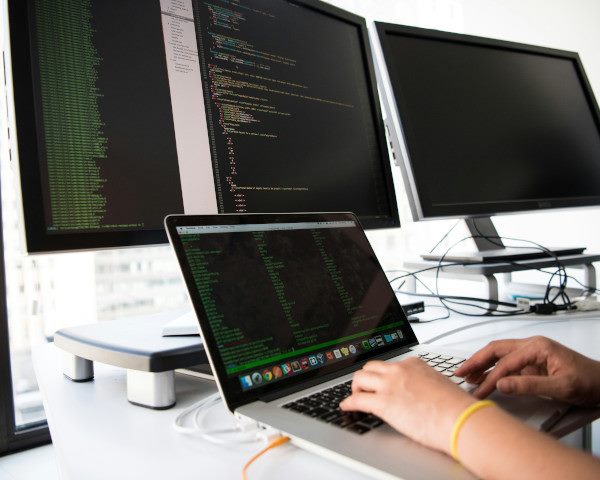 An employee writes code on a laptop computer with two external monitors.