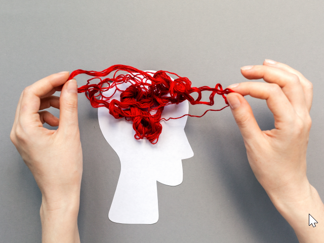 Female's hands unravel tangled red threads on the white paper silhouette of a head, representing the brain. on a gray background and lying flat.