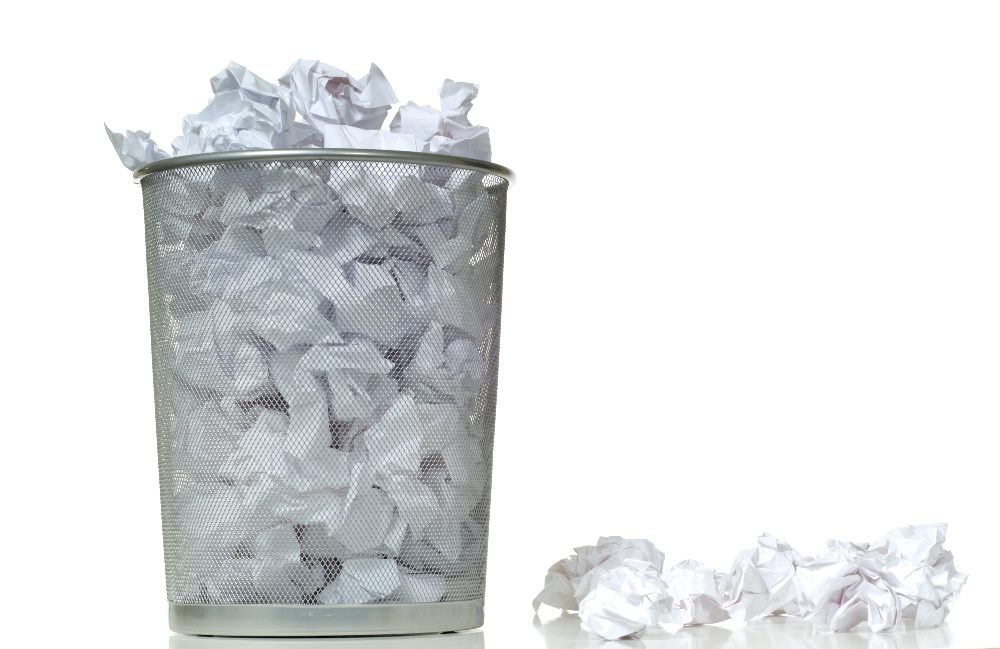 A mesh trash can is filled with wads of paper and is overflowing suggesting the need or a trial-and-error approach for AI in business.