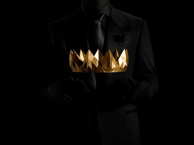 gold crown being held by a figure in a black bodysuit, with a black suit on top