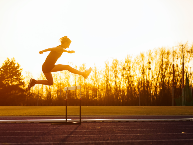 An athletic woman jumping above the hurdle on stadium running track during sunset