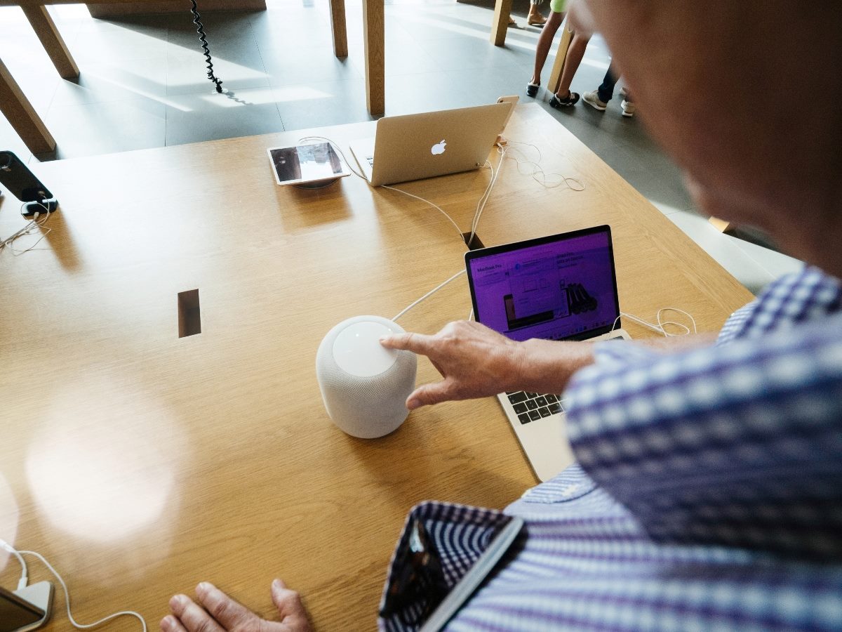 A person taps the top of a white Apple HomePod smart speaker on a wooden table inside an Apple Store, with MacBooks and other devices displayed nearby.
