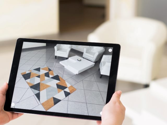 A customer holds a tablet up in a room to preview a carpet that has geometric black, gold, gray and white triable design. 
