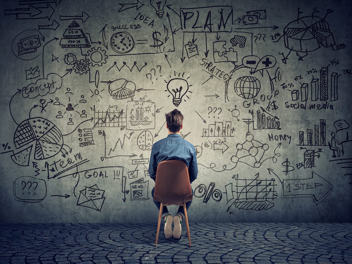A man sitting in a chair facing a wall, the wall covered with doodles of charts, squiggles, lightbulbs and more