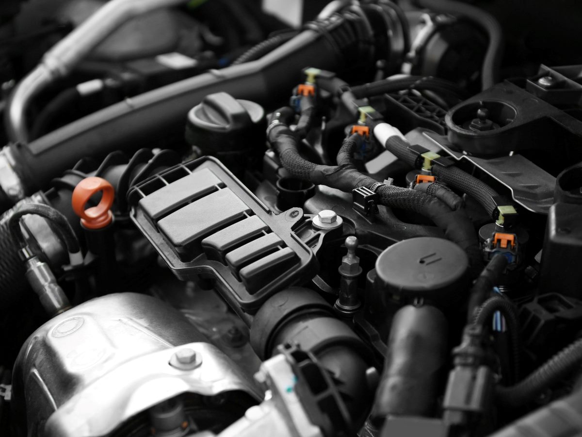 Close-up view of a modern car engine bay, dominated by black plastic engine covers, molded cable conduits and a dense network of wiring harnesses with color-coded orange and yellow connector clips running across the top of the motor. A silver metallic intake or turbo housing sits in the lower left foreground with a visible bolt head, while an orange-handled dipstick loop and a white-capped fluid reservoir punch through the otherwise monochrome gray and black palette of hoses, brackets and fasteners. The shot is tightly framed with shallow depth of field, blurring the surrounding hoses and firewall components in the upper corners to keep focus on the central cluster of connectors and covers.