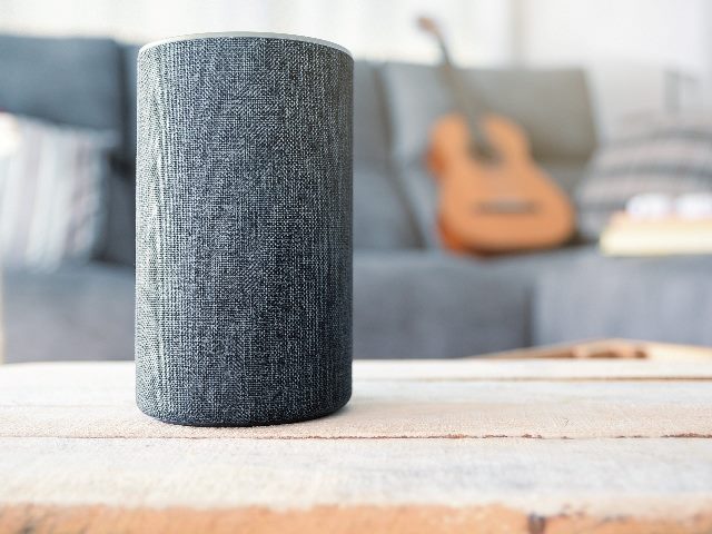 Personal assistant connected loudspeaker on a wooden table in a Smart Home in a living room. A guitar and some books on a sofa in the background.
