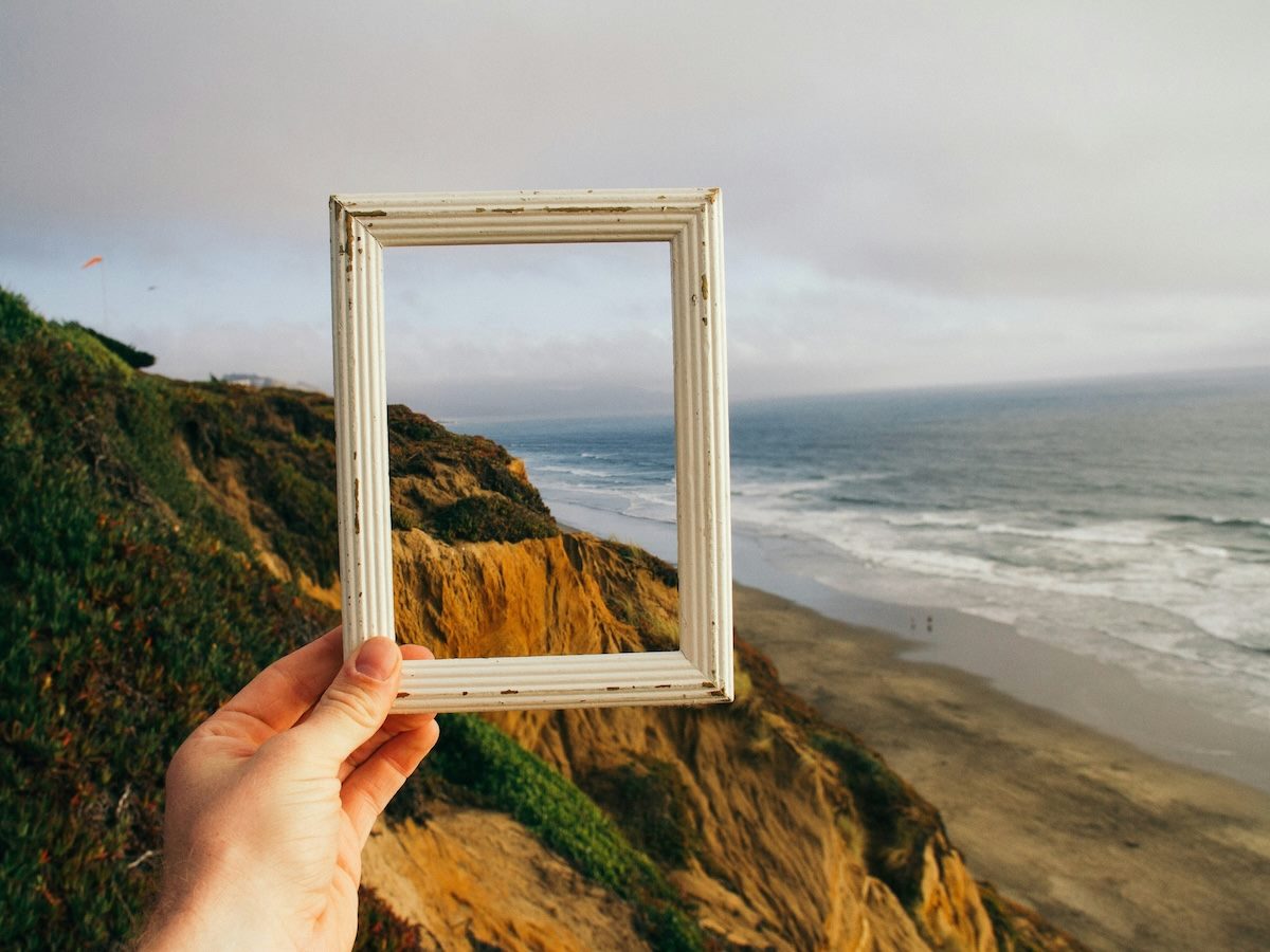 a hand holding up an empty picture frame. In the background, a cliff and ocean landscape