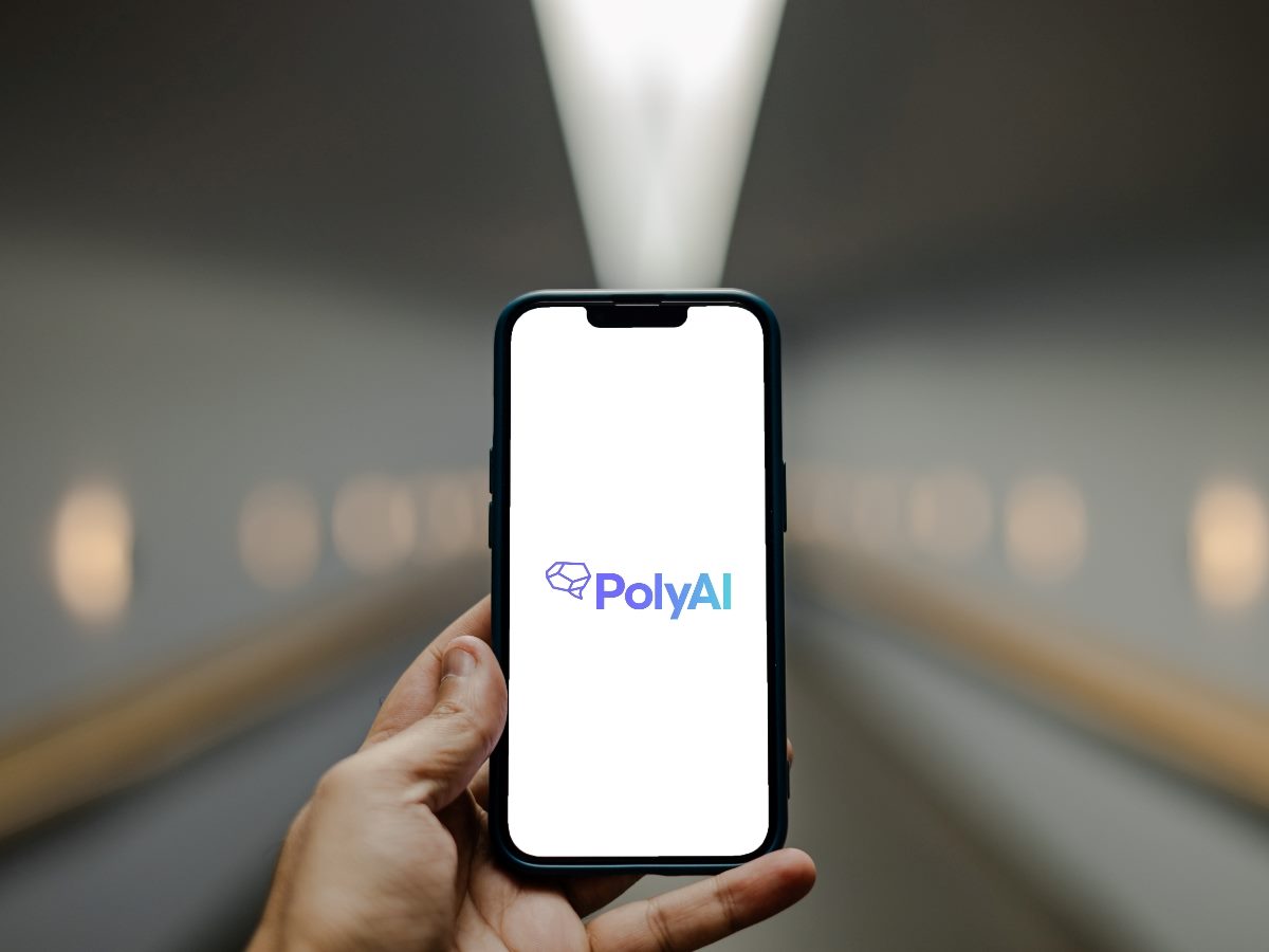 A hand holds a smartphone displaying the PolyAI logo against a blurred corridor background with overhead lighting.