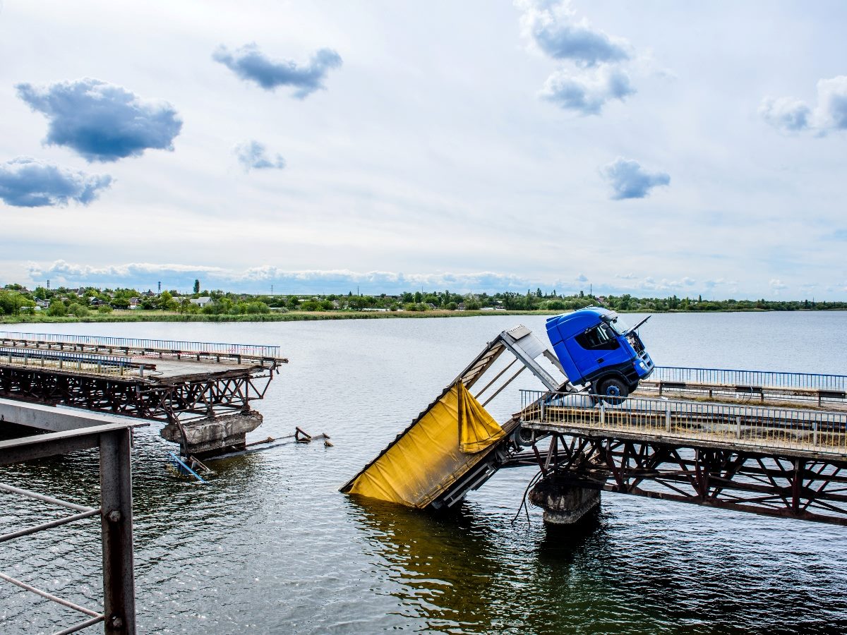 A blue semi-truck sits precariously on the broken edge of a collapsed bridge spanning a body of water. A large section of the bridge has fallen into the water below, leaving a wide gap between the remaining sections. The scene is set under a cloudy sky, with shoreline vegetation and distant buildings visible in the background.
