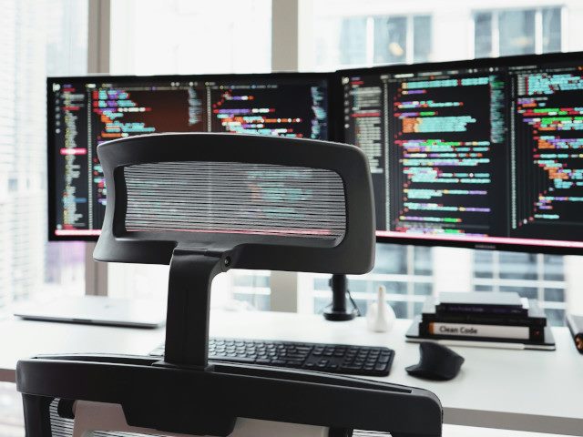 Two external computer monitors on a desk next to a window in an office building.