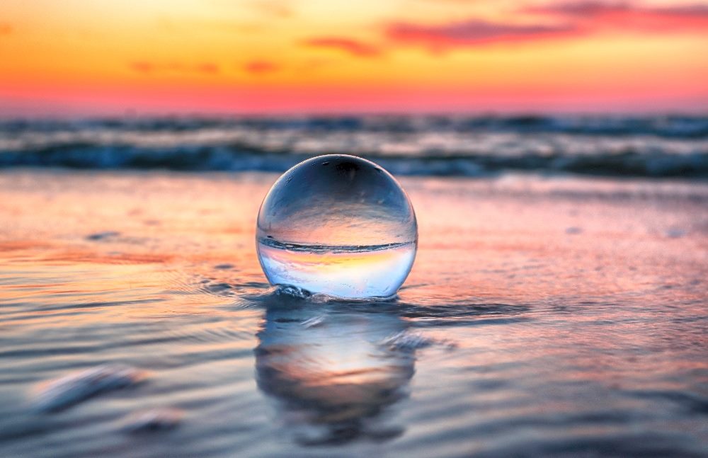 A crystal. ball sits on a beach with waters lapping around it and a gorgeous sunset in the back ground in Slowinski National Park near Leba, Poland in piece about predictive analytics in CX.