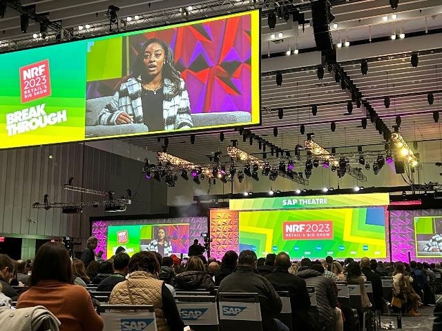Inside a keynote conference room at NRF 2023 conference in New York City. A big monitor hangs from the ceiling with a woman speaking as attendees listen and sit in chairs.