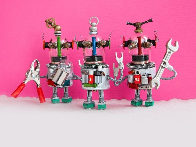 Three robots made from metal scraps hold a set of pliers, a light bulb and a wrench against a hot pink and white background in piece about AI content creation tools. 
