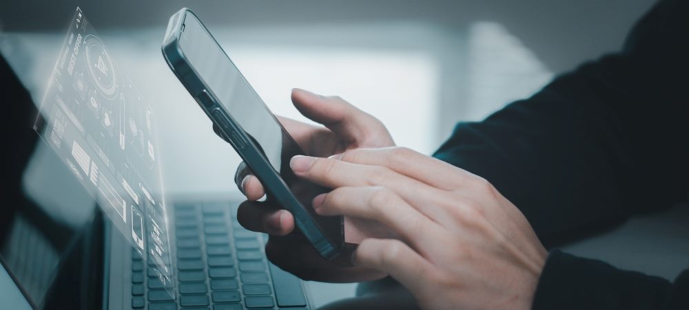 A man's hands using a mobile smart phone with a laptop visible in the background, representing the use of AI in omnichannel strategies.