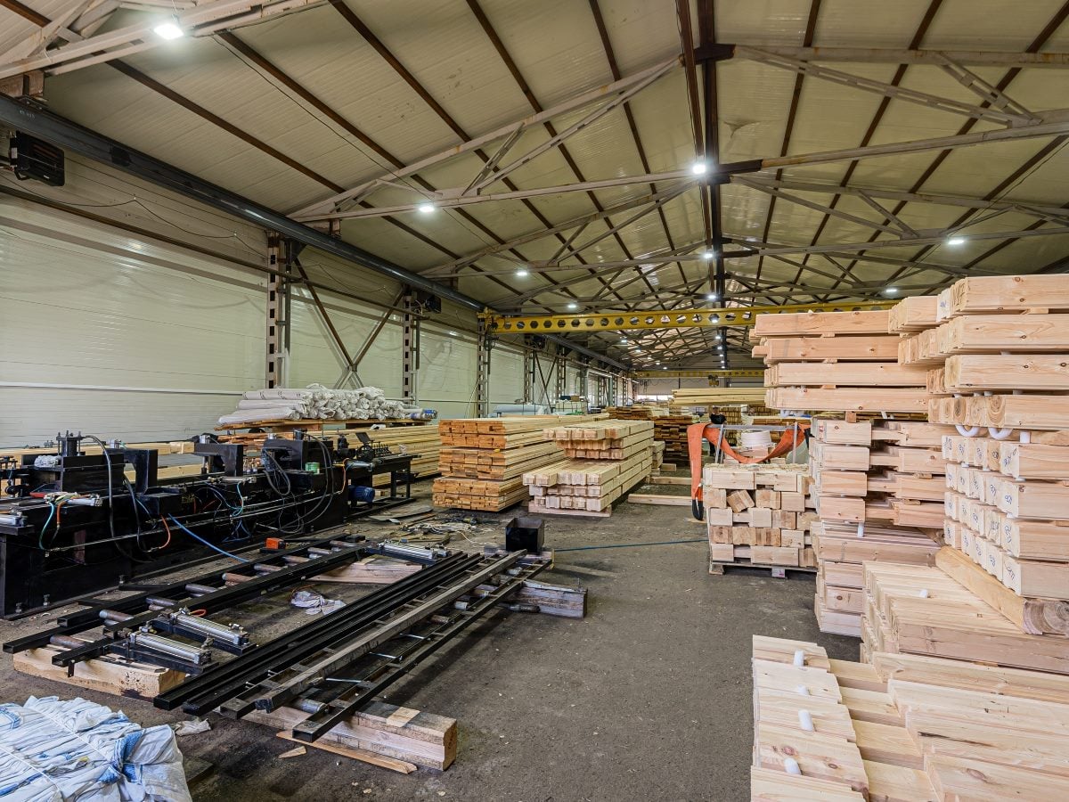 Industrial warehouse interior with large stacks of lumber and cut wood beams on pallets beside woodworking machinery under a high, metal-framed ceiling with overhead lights.