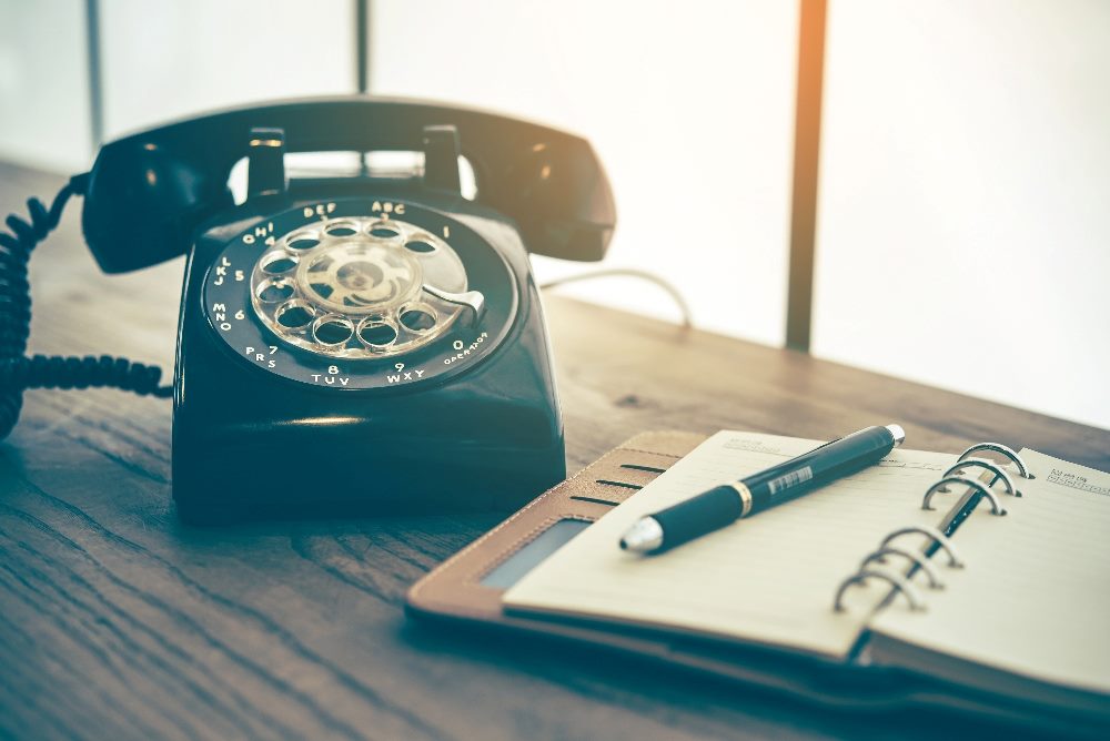 Alternate text description: A retro black rotary telephone sits on a wooden desk, accompanied by an open notebook and a pen, highlighting traditional communication tools contrasted with the advanced capabilities of AI in customer service. The scene is bathed in warm, natural light, evoking a sense of nostalgia yet emphasizing the evolution to modern customer service technologies.